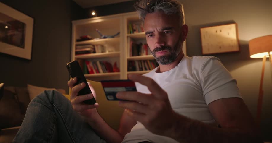 A man sits on a couch holding a credit card in one hand while making a purchase on his smartphone with the other