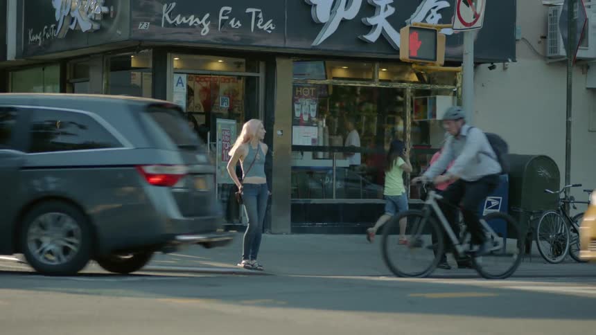 A female pedestrian stands on a city street corner as traffic and people flow around her