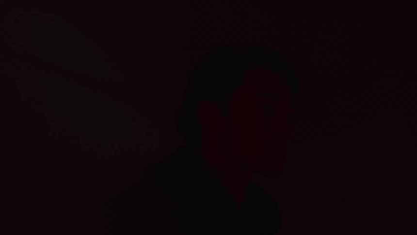 A young man in a jacket stands in a dimly lit room illuminated by flashing blue and red lights