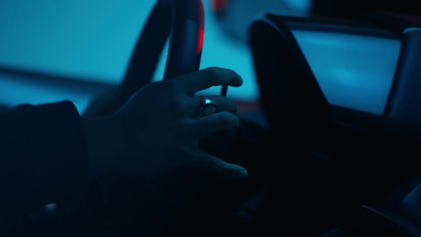 A person sits in the front seat of a car holding the steering wheel as the garage lights flicker between red and blue