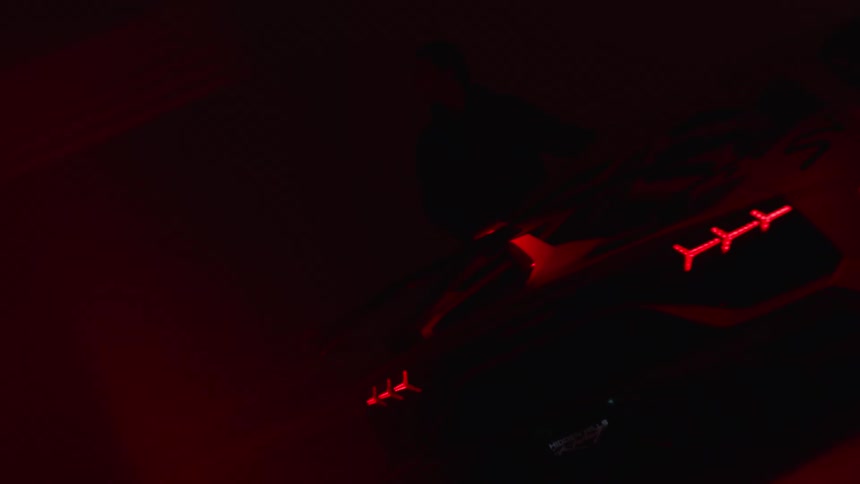 A young man in a jacket stands in a dimly lit room next to a modern sports car illuminated by flashing blue and red lights