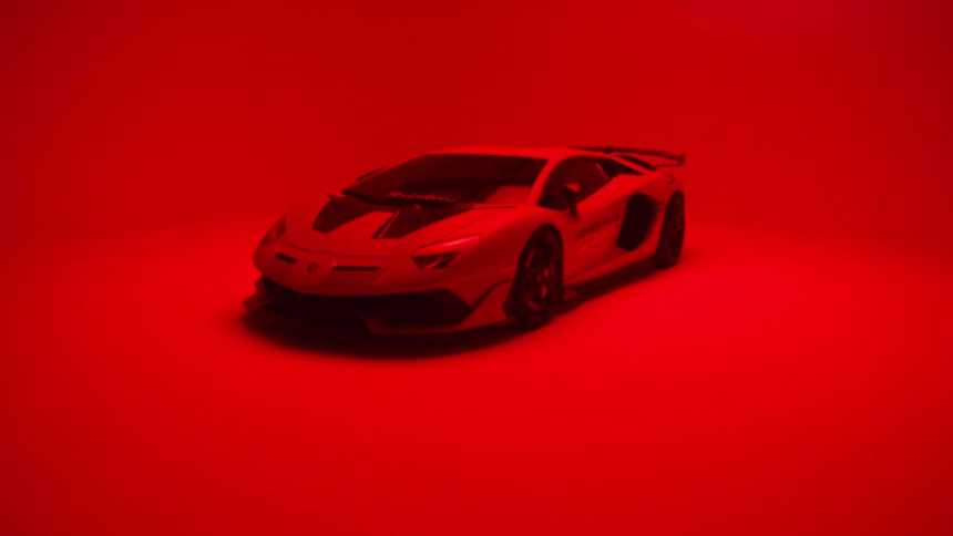 A white lamborghini aventador svj with black racing stripes is bathed in intense red light against a neutral backdrop