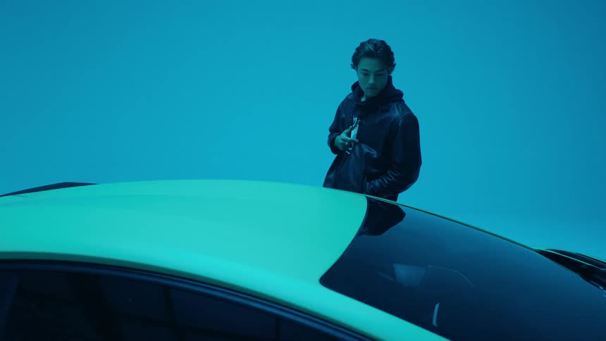 A young man in a jacket stands next to a car in a dimly lit room illuminated by flashing blue and red lights