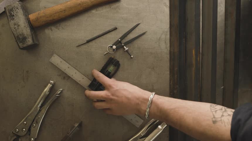 Mans hand with a tattoo and bracelet picks up tools on a metal workbench
