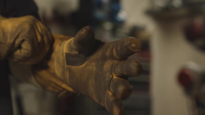 A man picks up his gloves puts them on then grabs a welding tool and begins welding iron inside the workshop