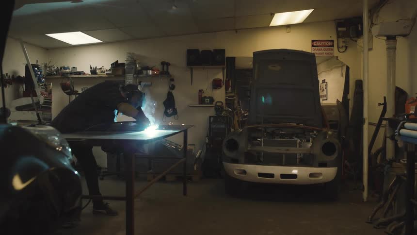 In a cluttered garage a person wearing a welding mask is working on a metal surface with a partially disassembled car nearby