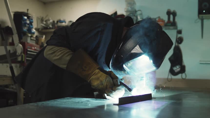 Welder works concentrated on a metal piece with sparks and intense light illuminating the environment