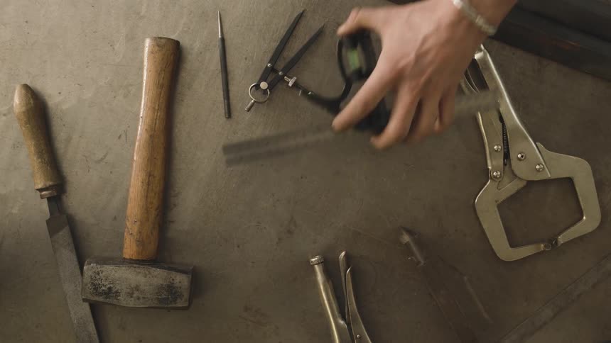A hand with a bracelet picks up a ruler and level among other tools on a gray surface