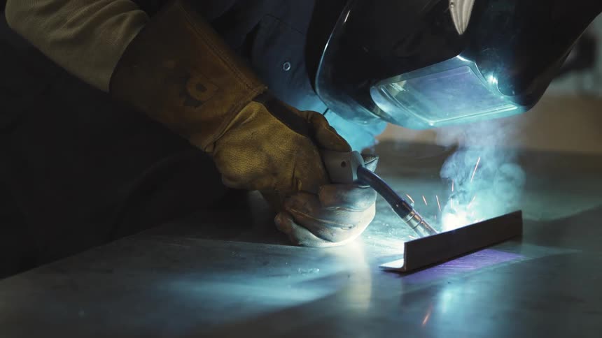 A person wearing a protective helmet is welding a piece of metal and releasing several bright blue sparks
