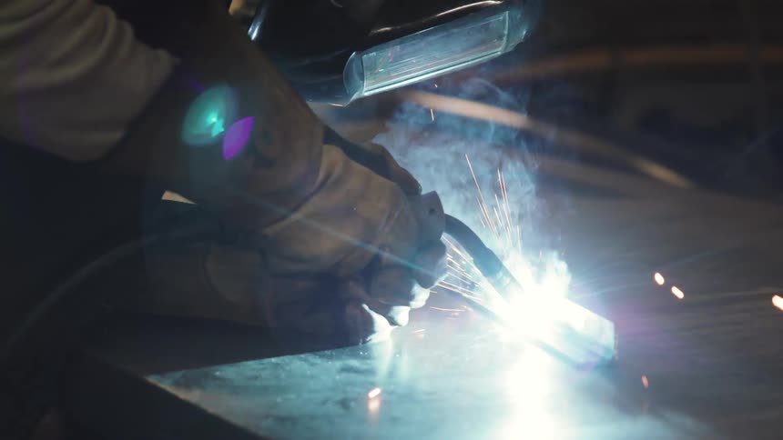 A person wearing a protective helmet is welding a metal object releasing several bright blue sparks
