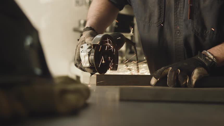 Gloved hands guide a spinning angle grinder across a metal surface producing a spray of sparks