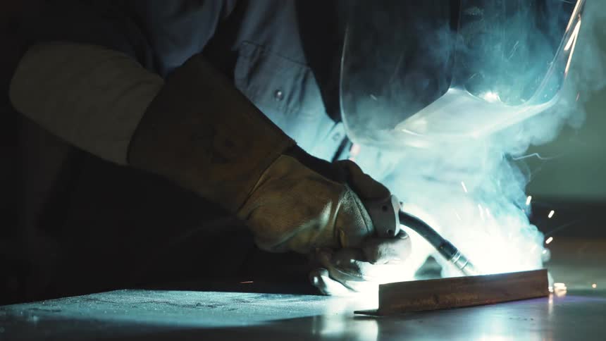 A person wearing protective gloves welding an iron object and releasing several bright blue sparks that are reflecting on his helmet