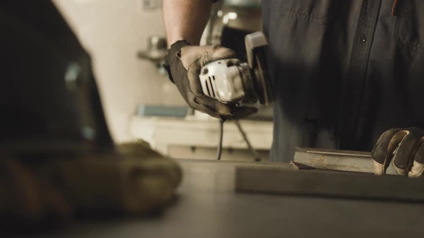 A person wearing protective gloves is using a machine with a rotating disc to cut a piece of metal and releasing several bright sparks