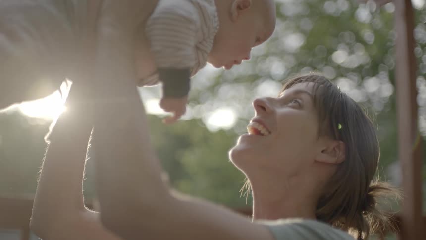 A woman holding a baby up in the air outside and smiling at it