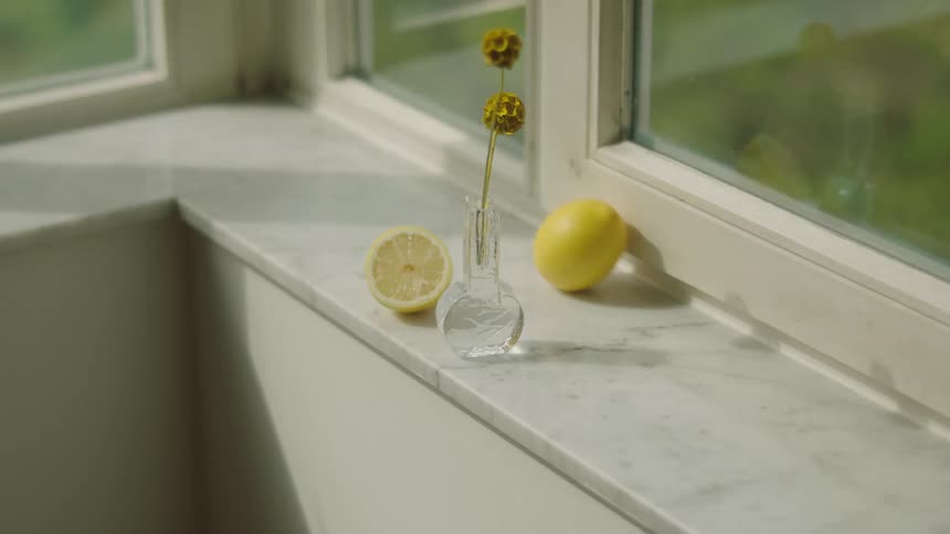 A lemon rolling by a vase on a marble window sill