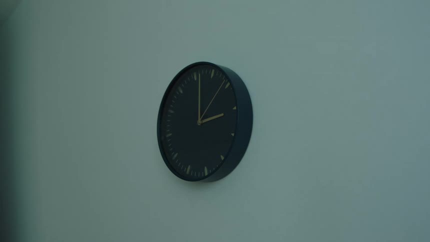 A dark wall clock hangs on a pale wall its hands pointing to 1200 under soft indoor lighting