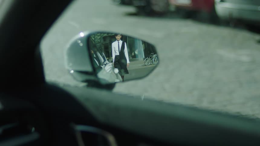 A woman in formal wear is seen walking toward a car reflected in its side mirror on a city street