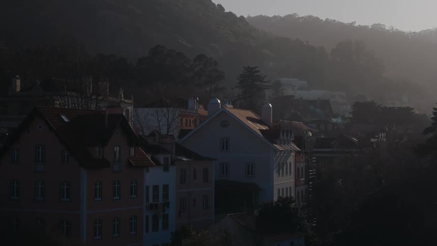A quiet picturesque town sits at the base of a forested mountain bathed in soft dawn or dusk light