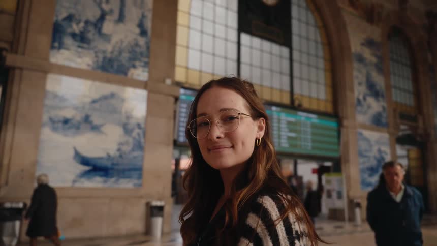 A young woman smiles inside a historic train station with tiled murals and travelers in the background