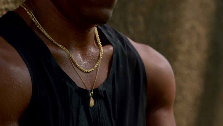 A black man stands beside a wall wearing a black tank top and two gold necklaces stretching his shoulder and smiling