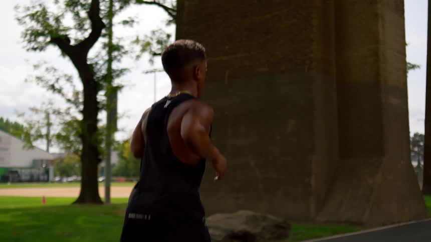 A man runs confidently along a park trail flanked by tall stone pillars and trees on a bright day