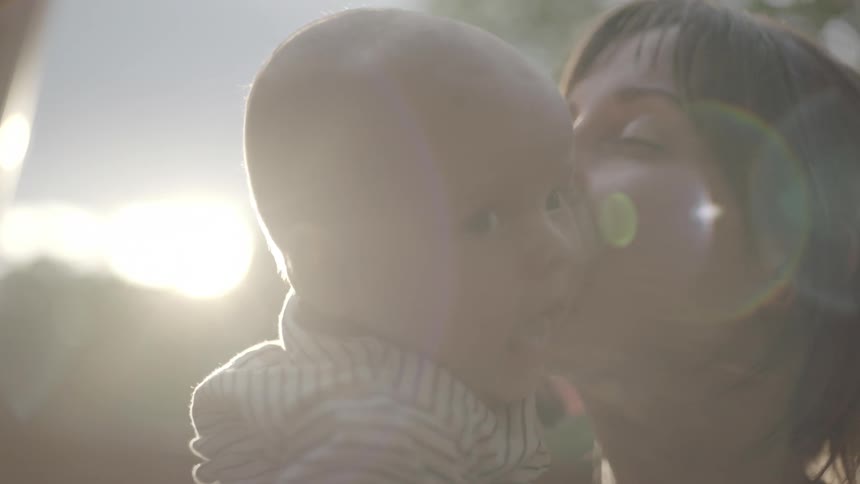 A woman holds and kisses a baby with the sun shining behind them