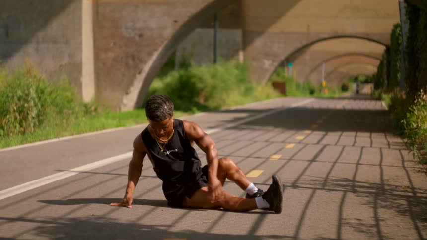 A young man rests on a sunlit path after an intense workout beneath a series of arched bridges