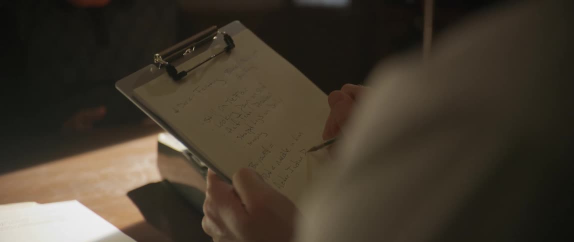 A person holds a clipboard with handwritten notes over a wooden surface with scattered papers