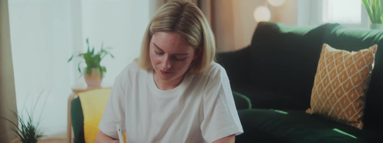 A woman sits on the floor beside a coffee table writing on a tablet with a pen at midday