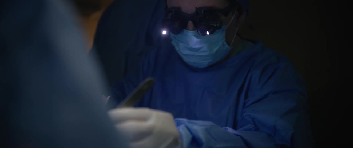 A focused surgeon operates under bright lights using magnifying loupes