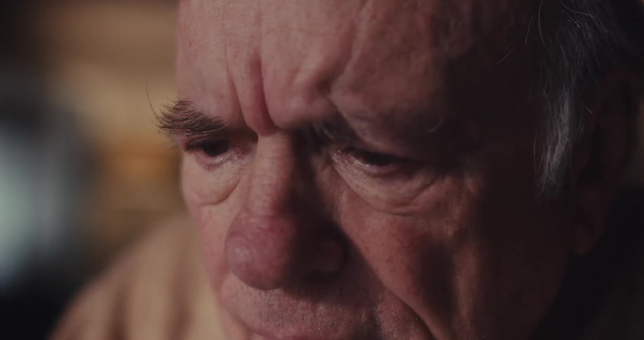 An elderly man wearing a sweatshirt sits indoors looks at a computer screen and starts smiling