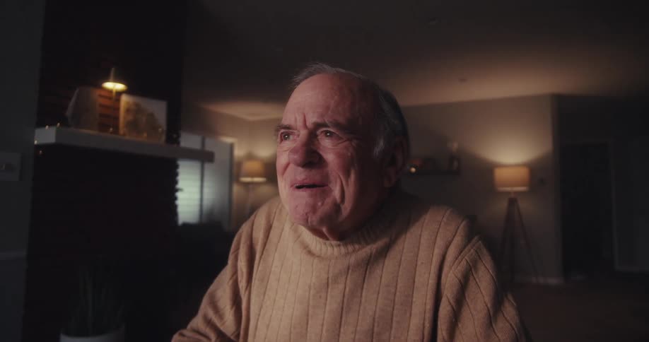 An elderly man looks concerned during a video call in a dimly lit room