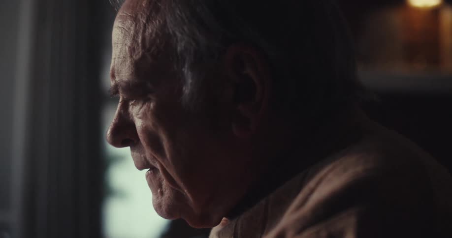 An elderly man sits near a window thoughtfully smiling during a video call in a warmly lit room
