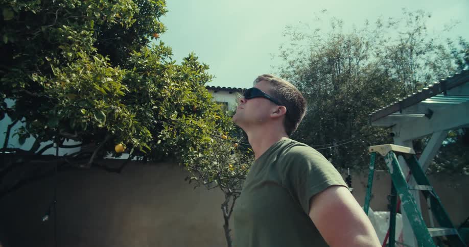 The camera moves from behind to front of a man in sunglasses capturing his intense upward gaze in a sunlit backyard