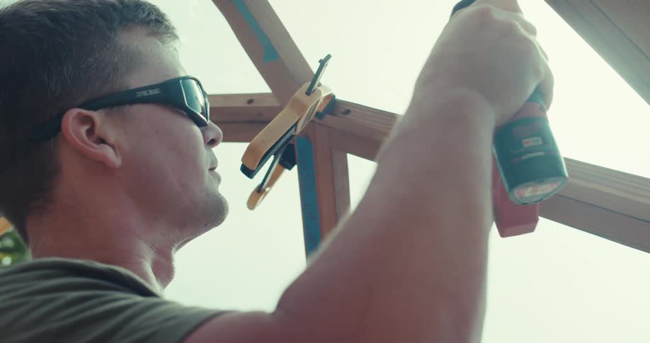 Man in sunglasses drills wooden beams under bright daylight