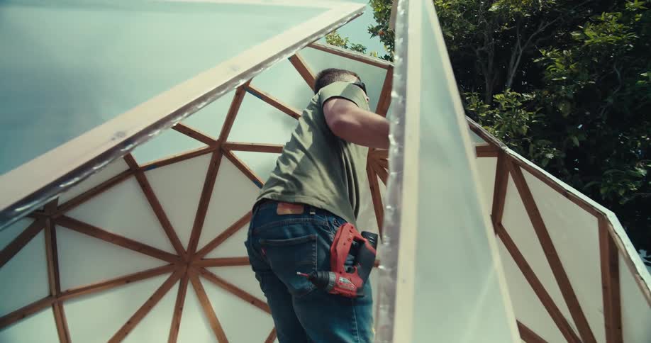 Man in sunglasses builds a geodesic dome with wooden beams in daylight