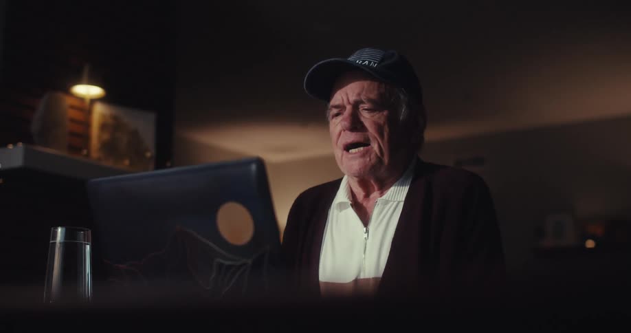 An elderly man in a cap video chats at night focused on his laptop in a dimly lit room