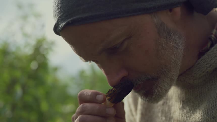 A man intently smells a mushroom in the outdoors hinting at foraging or culinary curiosity