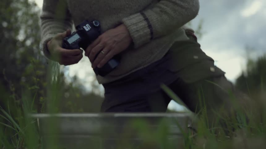 A man in a knit sweater crouches in grass photographing something in a forest