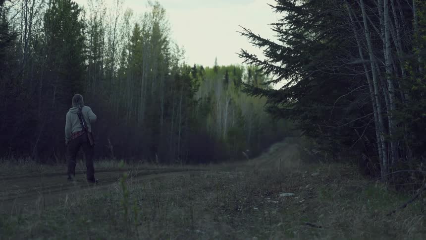 A hunter carries a rifle while walking down a dirt road in an evergreen forest