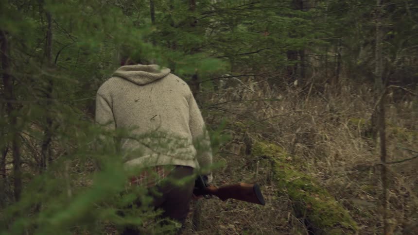 Back of a man holding a shot gun walking through trees in the forest