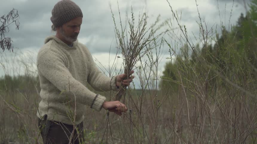 A man in a knit sweater and beanie cuts wild plants with garden shears in a dry open field