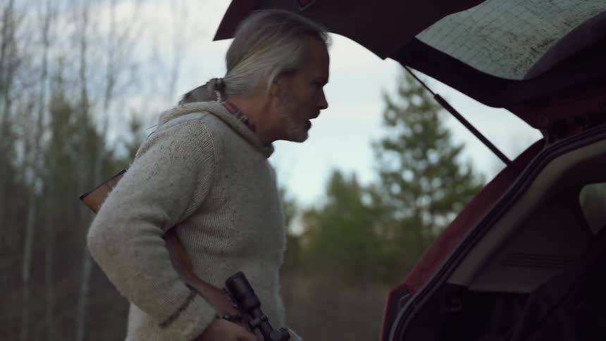 A man with a rifle places a hunted rabbit into the back of a vehicle in a forest at dusk