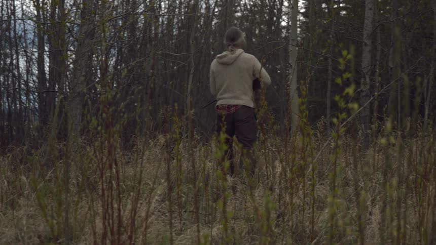 A person carrying a rifle walks through a grassy field near a tree line under a cloudy sky focused on hunting