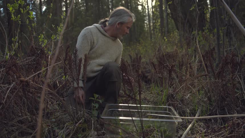 A man crouches in a forest examining a plastic tub on the ground