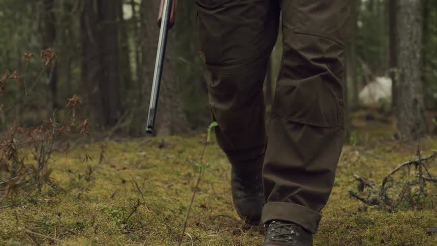 Close up of mans boots walking on dried brush through the forest carrying a rifle