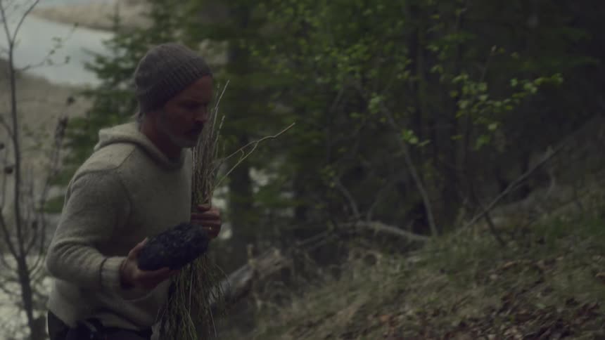 A man walks uphill in the forest at midday holding a bundle of grasses and a rock surrounded by trees