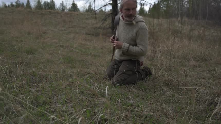 A hunter kneels in the grass carrying a rifle over his shoulder while following tracks
