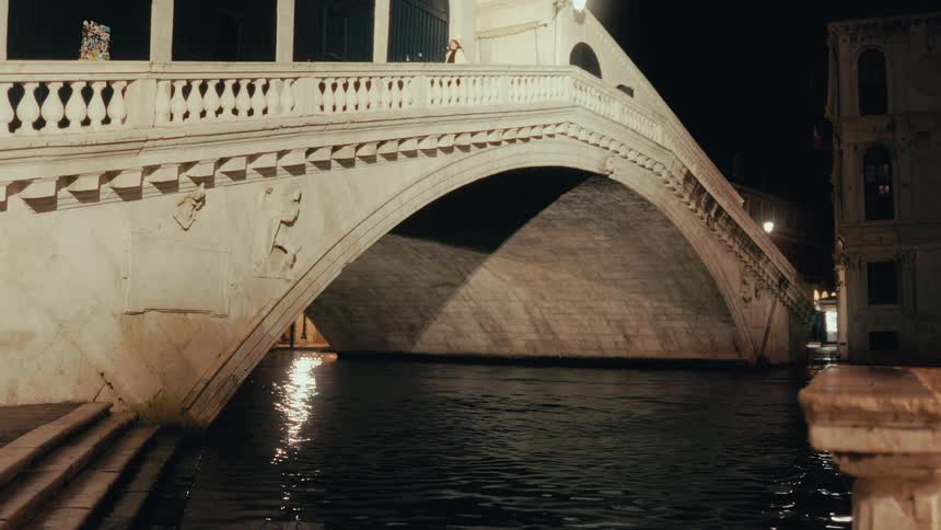 An ornate stone bridge illuminated at night arches gracefully over a dark canal as people stroll across its walkway