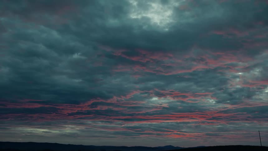 A wide shot captures a dramatic sunset sky filled with dark clouds tinged with red set above a silhouetted landscape of hills